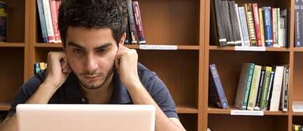Ein junger Mann liest konzentriert in seinem Laptop. Im Hintergrund steht ein Bücherregal.