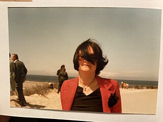 Eine Fotografie, auf einer Unterlage liegend, zeigt eine Frau mit getönter Brille, eine Haarsträne verdeckt einen Teil ihres Gesichts; im Hintergrund Menschen, Strand, Meer. © Foto: Nora Gomringer Den Tod der Mutter beschreiben