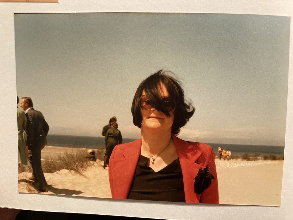 Een foto, liggend op een onderlegger, toont een vrouw met getinte bril, een lok haar bedekt een deel van haar gezicht; op de achtergrond, mensen, strand, zee.
