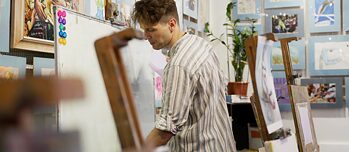 Ein Mann schreibt auf dem Whiteboard, im Hintergrund ist Kunstwerkstatt mit Gemälden