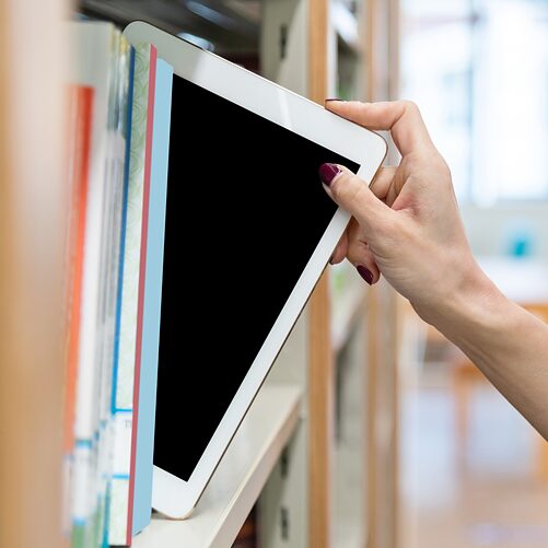 Eine Frauenhand nimmt ein digitales Tablet aus dem Bücherregal einer Bibliothek. Bildauswahl zur Kultur- und Spracharbeit.