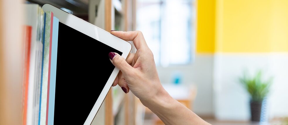 Eine Frauenhand nimmt ein digitales Tablet aus dem Bücherregal einer Bibliothek. Bildauswahl zur Kultur- und Spracharbeit.