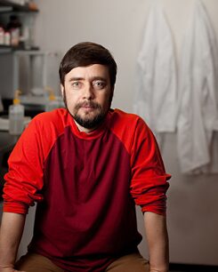 Mann sitzt mit rotem Pullover in einem Labor mit hinten hängenden Laborjacken