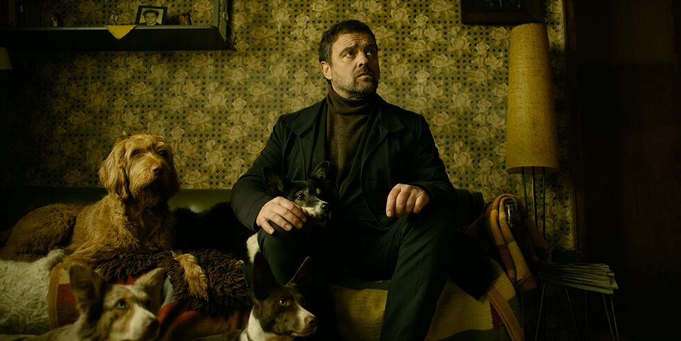 Still frame from the ZDF Series "Perfume" - "Perfume - Ambra": Main Commissioner Matthias Köhler (Jürgen Maurer) is sitting on a couch. Next to him, as well as in front of him, there are two dogs sitting.
