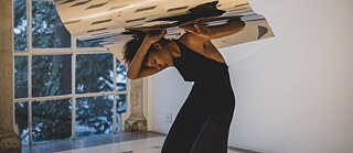 Performance im Rahmen der Projekte "Halaquat" und IDEA-Festival Eine tanzende Frau in schwarzem Kleid hält mit beiden Händen einen gewellten Spiegel über ihren Körper.