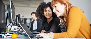 Zwei lachende, junge Frauen sitzen in einem Büro am Schreibtisch. Vor ihnen stehen mehrere Bildschirme und ein Laptop.