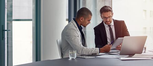 Zwei Männer sitzen in einem Büro und schauen zusammen ein Papierdokument an, vor ihnen steht ein Laptop