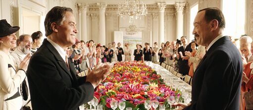 In an elegant white hall with high ceilings there is a table set with festive flowers. Dishes, wine glasses and cutlery is set on the table. Well-dressed people in old fashioned attire of the era of the Second World War are standing around the table. They are applauding and looking at the two men in the front, who are dressed in suits, looking at one another and are smiling.