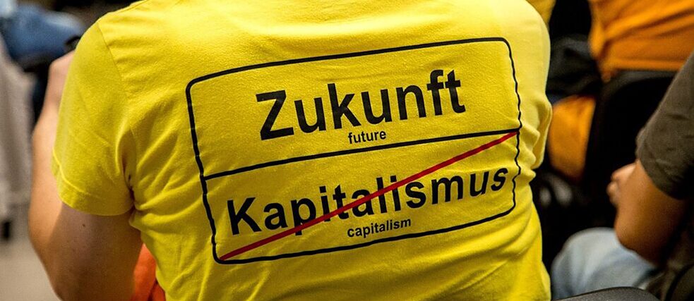 Una persona che partecipa a un convegno indossa una maglietta con la scritta “Futuro | Capitalismo (barrato)” nello stile di un segnale di uscita dalla città.