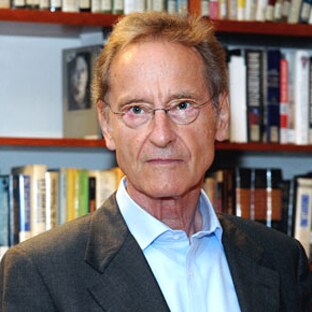 Bernhard Schlink is standing in front of a bookshelf. He is wearing a light coloured shirt, a dark coloured blazer and glasses. 