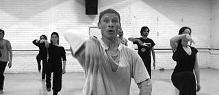 The black and white image shows a group of dancers in a dance studio. The people are all doing the same dance movement. In the foreground is a dancer in a white t-shirt, all the others are dressed in black.