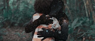In the background of the picture is a forest. In the front you see a child from behind. This child wears a striped top and has afro hair. It is embraced by a black human silhouette, on which galaxies are depicted.