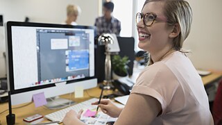 Blonde Frau sitzt vor einem Computerbildschirm und schaut lachend zu eine Kollegen auf