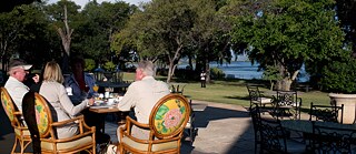 Several White people are sitting on a hotel terrace, in the background you can see a garden and water, further back a Black person is standing