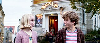 The picture shows two children in a semi-close-up shot, who are talking to each other on a busy square, looking at each other. The boy, on the right, with brown short curls, laughs lively. The girl, left, light blond with chin-length hair looks at him interested and friendly.