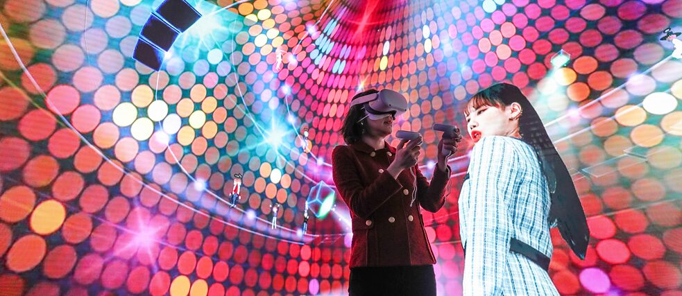 Two women in front of a colourful background one with VR glasses 