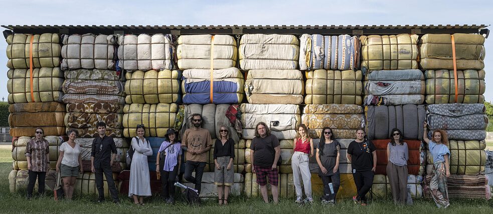 A group of 13 people are standing in a row on a grass field. Behind them is a wall made out of folded matresses stacked one on top of the other.