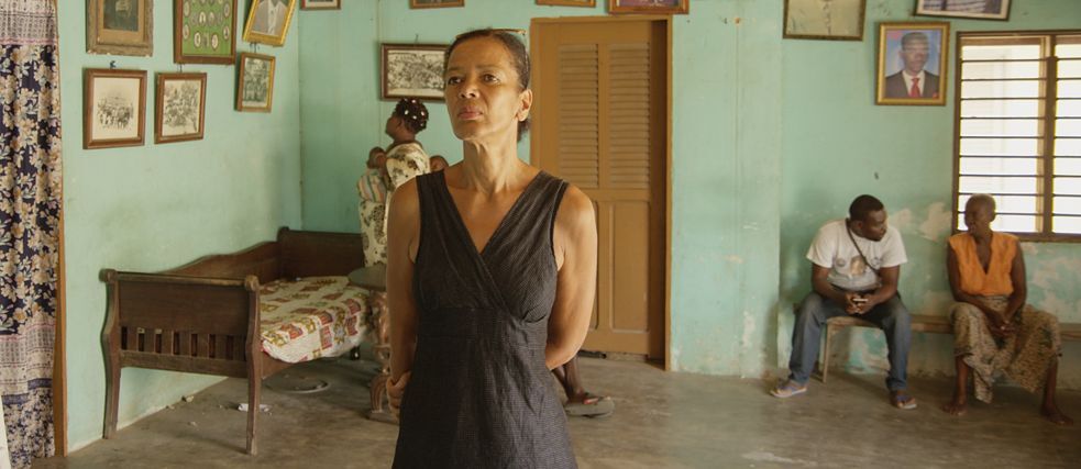 Becoming Black Foto eines Wartezimmers. Einige Menschen sitzen auf einer Bank. Eine stehende Frau im Vordergrund. Alle scheinen afrikanisch zu sein.