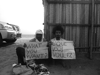 Two men sit in the desert with signs that say "what you want?". 