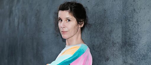 Die Autorin Daniela Dröscher steht mit verschränkten Armen vor einer Betonwand. Sie hat braune, lockige Haare, die im Nacken zusammengebunden sind und trägt einen Pullover mit geometrischen Mustern in  hellblau, türkis, orange und rosa.