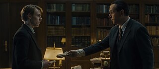 A young man holding a document is looking with a serious look at a middle-aged man, who is handing him another document. They are standing on either side of a desk with papers, a lamp and a telephone and in the background there are bookcases.