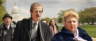 A tall man in a suit and a shorter woman in a blue coat are staring at something in surprise. Behind them there is a group of people and at the very back the White House can be seen.