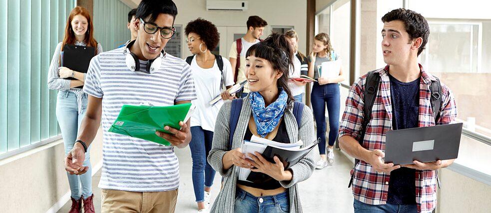 Mehrere Schüler laufen einen Korridor entlang und unterhalten sich dabei. Sie halten Unterrichts-Materialien und ein Laptop in der Hand.