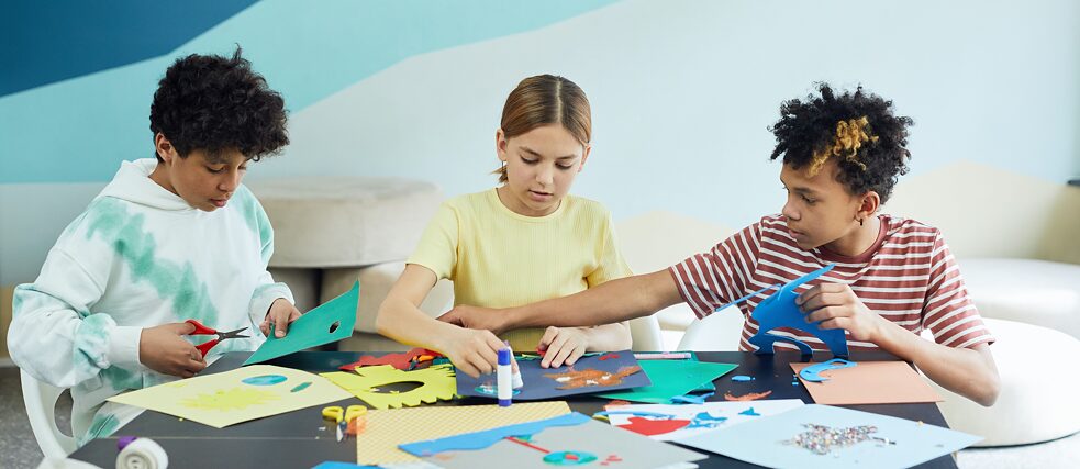 Zwei Jungs und ein Mädchen sitzen an einem Tisch und schneiden Muster aus Papier aus 