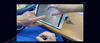 Ein Foto über einem dunklen Satellitenbild zeigt die Hände zweier Personen, die auf eine Karte zeigen, die auf einem Tablet-Bildschirm auf einem Tisch dargestellt ist. Die Untertitel lauten „Bewegen Sie es dort etwas mehr“.
