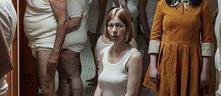 Szene aus dem Film 'The Ordinaries' von Sophie Linnenbaum. Ein Mädchen steht als einzig voll Bekleidete in einer Umkleidekabine mit halb angezogenen Erwachsenen