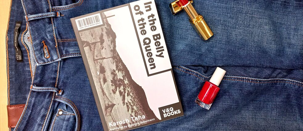 Das Buch 'In the Belly of the Queen' liegt mit rotem Lippenstift und Nagellack auf einer Jeanshose