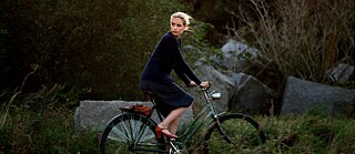 Scene from the film "Barbara": A woman with blonde hair wearing a blue dress is riding a bicycle in a green landscape, while looking over her shoulder.