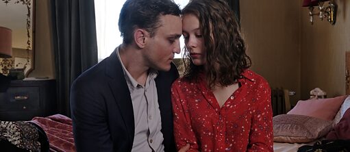 Scene from the film "Transit": A man in a suit and a woman in a red blouse are seated next to one another on a bed and facing each other. The room in the background appears to be a hotel room.