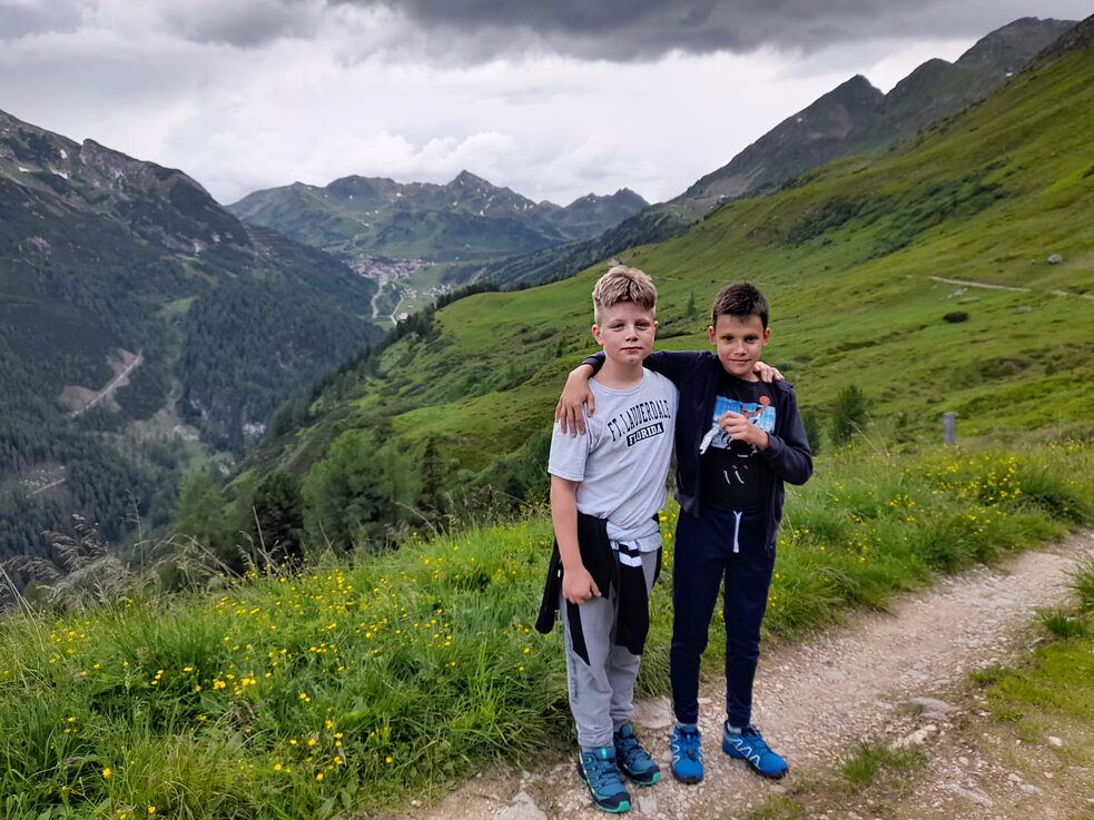 Zwei Jungs umarmen sich, im Hintergrund sind Berge und dunkle Wolken am Himmel.