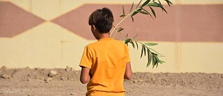  Ein Junge blickt zu einer braun-beigen Wand. Er trägt einen jungen Baum.