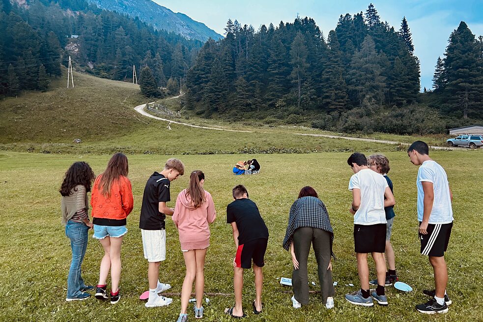 Jugendliche auf einer Wiese stehen im Halbkreis und blicken nach unten auf Notizen. Im Hintergrund Berge und Wälder.