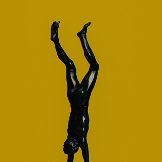 Siyah bronz bir erkek heykeli, sarı bir fon önünde amuda kalkıyor.