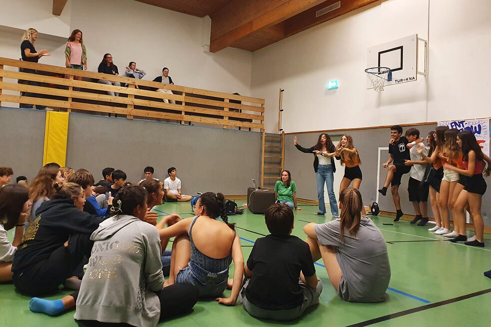 Jugendliche in einer Turnhalle sitzen auf dem Fußboden, andere stehen und singen.