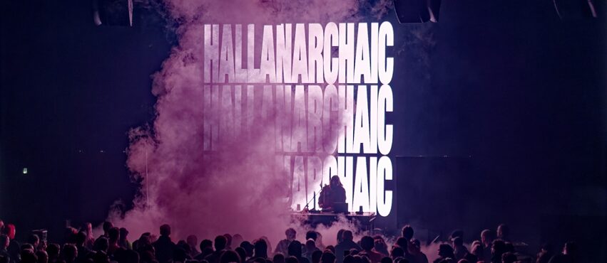 A stage with purple smoke in front of the crowd and HALLANARCHAIC printed on screen behind DJ