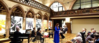 Foto eines Konzerts einer Solistin in einem blauen Kleid, begleitet von sechs Instrumentalisten und einem Dirigenten, in einem Saal mit gotischer Architektur, mit Kunstwerken im Hintergrund