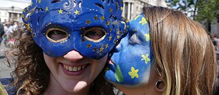 Eine Frau hat die Europaflagge ins Gesicht gemalt und küsst eine andere Frau mit Europa-Maske auf die Wange.