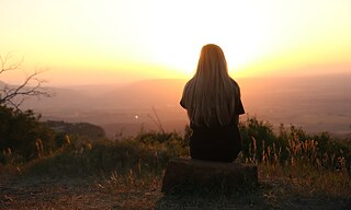 Eine junge Frau sitzt an einem Aussichtspunkt und sieht sich den Sonnenuntergang an. 