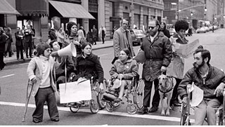 影片中的残障者在城市街头游行，他们之中有的手拄拐杖，有的坐着轮椅，有的牵着导盲犬