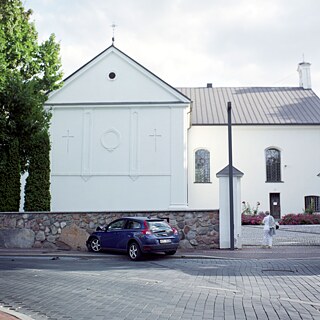 Dieses Foto hält einen außergewöhnlichen Moment fest - ein unvorsichtiges Auto kollidierte mit der Mauer der Basilika des Erzengels Michael. Die Basilika ist weiß, mit präzisen, sauberen Formen, einer bescheidenen Fassade und kleinen Kreuzen, die die Giebel zieren. Eine weiß gekleidete Frau betritt den Kirchhof, auf dem rote und violette Blumen blühen. Das kleine dunkelblaue Auto ist gegen die Mauer geprallt. Die Motorhaube ist verbogen, die vordere Stoßstange liegt unter den Rädern auf dem Bürgersteig. Dunkle Bremsspuren bleiben auf dem Kopfsteinpflaster zurück.