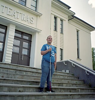 Das Farbfoto zeigt Mangirdas auf den Stufen des Dramatheaters von Marijampolė. Er trägt ein blaues T-Shirt mit weißer Schrift, blaue Jeans und dunkle Schuhe und hat eine goldene Kette um den Hals. Der Junge ist in ganzer Größe abgebildet und lächelt bescheiden, während er auf der grauen Betontreppe steht mit einem Blindenstock, der sich steil auf die Stufe unter seinen Füßen stützt. Mit der rechten Hand hält er das Ende des Stocks fest umklammert, die linke Hand hat er säuberlich auf den Oberschenkel gelegt. Hinter Mangirdas steht ein nüchternes weißes Gebäude mit einem auffälligen Schild über der Eingangstür, das darauf hinweist, dass es sich um ein Theater handelt; das einzige Wort, das es auf das Foto geschafft hat. Die Buchstaben sind unterschiedlich hoch und breit, die Schrift ist etwas unordentlich, sieht verspielt aus. Der Zugang zum Theater erfolgt durch eine braune, rechteckige Tür. Wenn man das Theater verlässt, muss man eine breite Treppe hinuntergehen, die an beiden Enden von dunklen Wänden und weißen Handläufen eingerahmt ist. An der Ecke des Gebäudes stehen grüne Bäume.