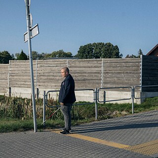 Dieses Farbfoto zeigt einen Straßenrand. Mangirdas, der auf einer gelben taktilen Linie den Bürgersteig entlangging, steht wie erstarrt am Ende der Linie neben einem Pfosten mit Verkehrsschildern. Dies ist das Ende des taktilen Weges, wo der Junge mit einem weißen Stock die Grasnarbe berührt. Er trägt eine dunkle Jacke mit dem Emblem des St. Cecilia-Gymnasiums in Marijampolė am linken Revers nahe am Herzen, und eine dunkelgraue Hose, die mit dem Asphalt korrespondiert, sowie schwarze Turnschuhe. Mangirdas ist nachdenklich, seine Augen und sein Mund sind leicht geöffnet, sein Kopf ist kahlgeschoren, seine Stirn glänzt im Abendsonnenlicht, seine Silhouette wirft einen langen Schatten.