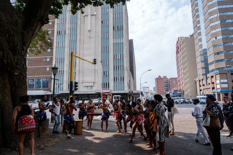 A performance from KCAP on the corner of Smith and Aliwal streets. The background features the church with brutalist architecture.