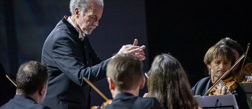 Eine kleine Gruppe Musikerinnen und Musiker spielen ihre Instrumente in einem Halbkreis und werden durch den Dirigenten angeleitet.