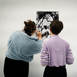 Двоє людей стоять перед картиною, яка висить на стіні. Одна з осіб тримає лінійку у руці.