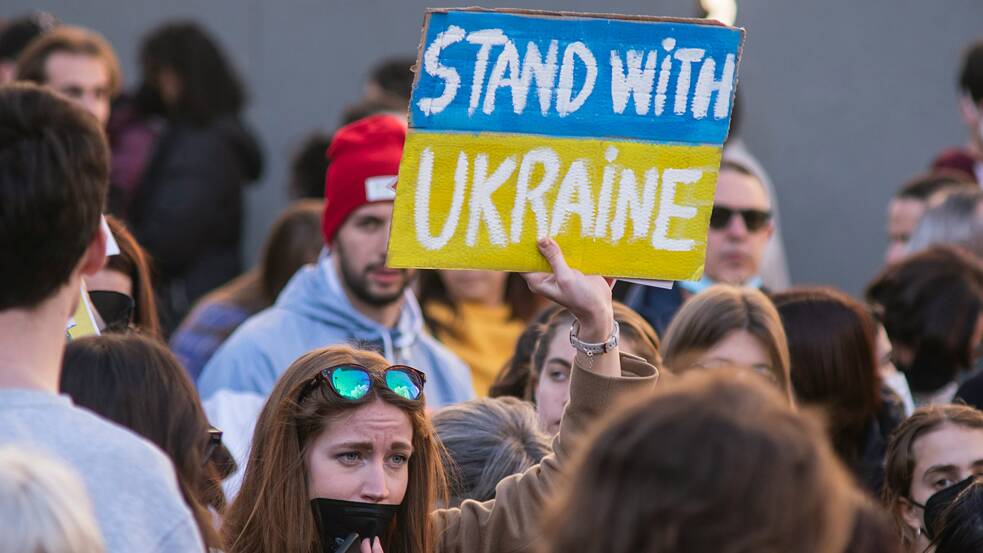 Photo is showing a demonstration with a woman holding up a sign that says “Stand with Ukraine”.